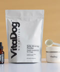 VitaDog nutrition products including a powder pack, dropper bottle, scooper, and a shaker on a beige background